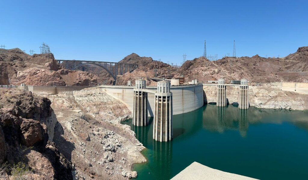 Wide view of Hoover Dam with intake towers and surrounding rocky landscape under a clear blue sky.
