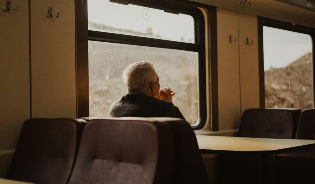 Elderly man sitting alone on a train, looking out the window with hands resting near his face.