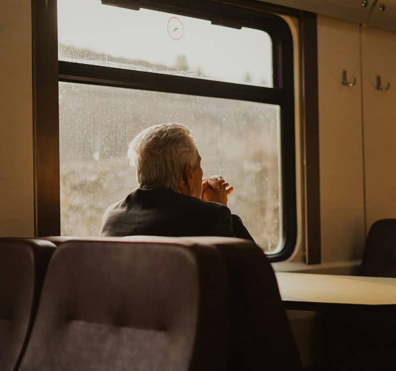 Elderly man sitting alone on a train, looking out the window with hands resting near his face.