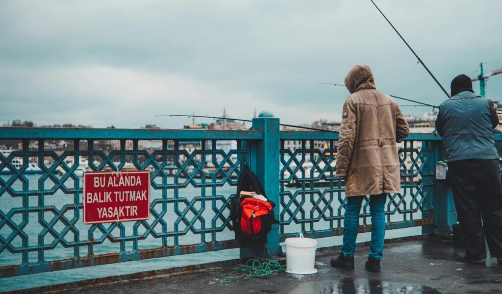 Two people fishing from a bridge with a "No Fishing" sign in Turkish attached to the railing, overlooking a body of water with boats in the background.