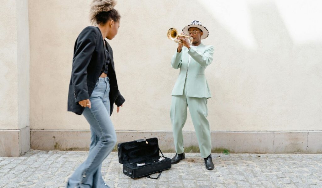 Street musician playing trumpet while a woman dances nearby, with an open instrument case on the ground containing money.