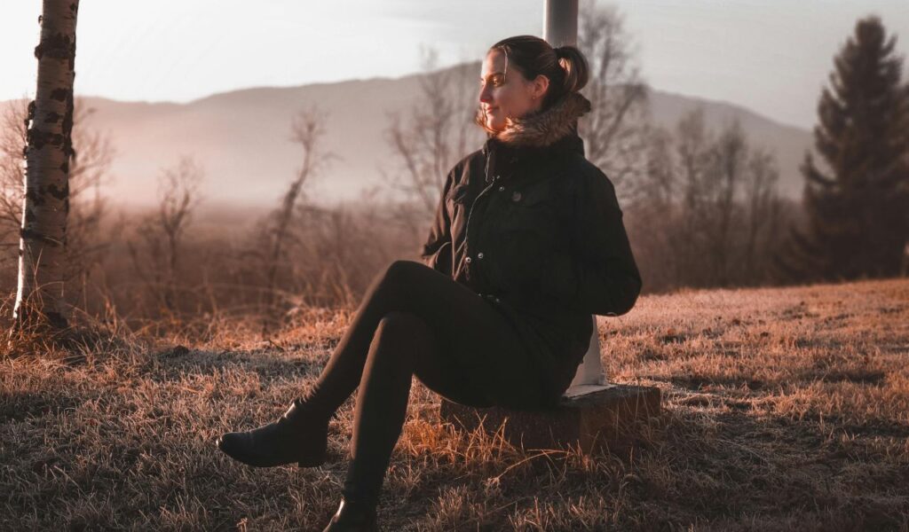 A woman in Norway sits on a low stone ledge in a frosty field at sunrise, dressed warmly in a dark coat with a fur-lined hood.