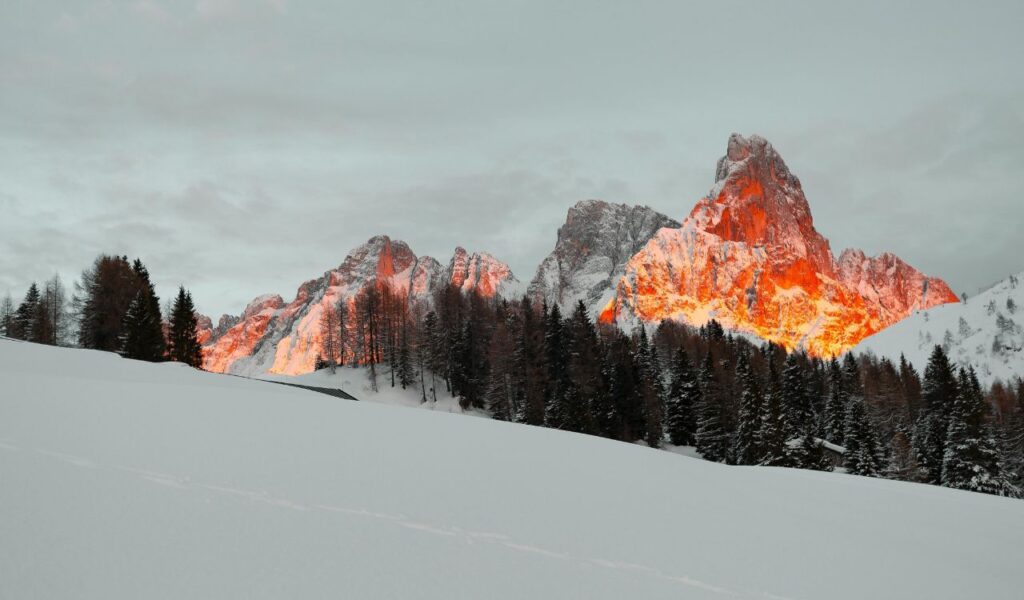 Snow-covered mountains and pine trees at sunset with fiery orange glow on rocky peaks in a quiet winter scene