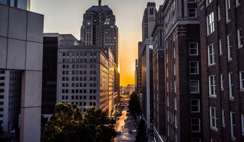 Street view of downtown Oklahoma City at sunset, with sunlight shining between tall buildings and trees lining the road.