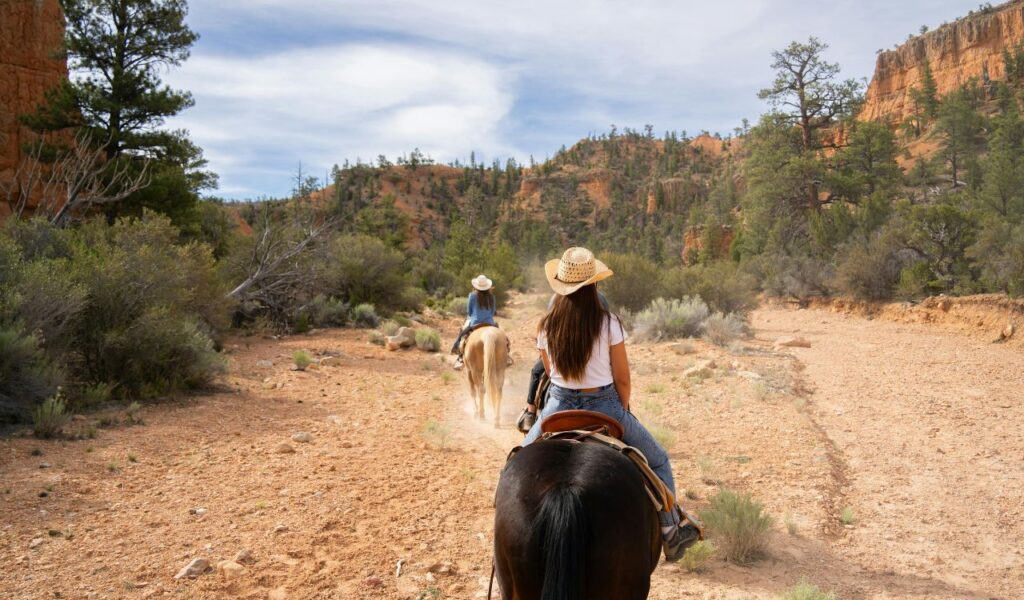 Two riders on horseback trail through the desert landscape of Onaqui Mountains, Utah, surrounded by rugged cliffs and sparse trees.