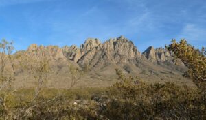 Rugged peaks of the Organ Mountains rise dramatically from the desert floor under a clear blue sky, framed by sparse desert vegetation.