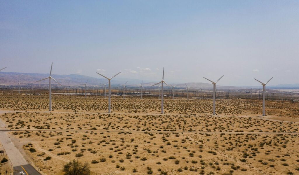Wind turbines in a desert landscape near Palm Springs, California, with scattered shrubs and mountains in the background under a hazy sky