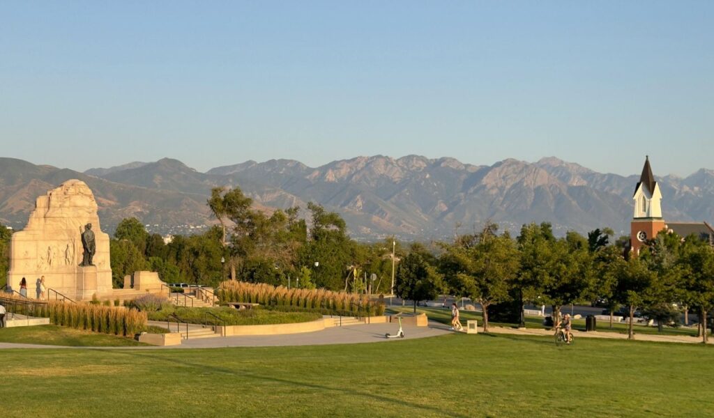 Scenic view from a park in Park City, Utah, with a monument, green lawns, and people walking, set against a backdrop of rugged mountain peaks under a clear sky.