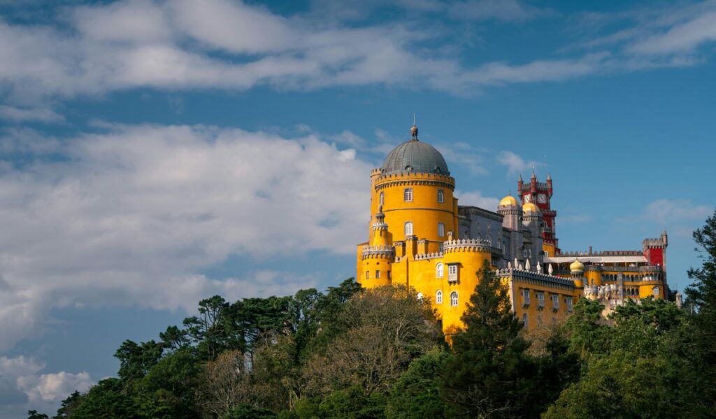 Pena Palace in Portugal with its striking yellow and red towers, domes, and ornate architecture, perched atop a forested hill under a partly cloudy sky.