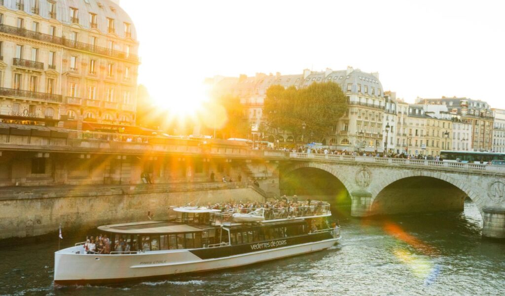 A modern river cruise boat filled with passengers sails along the Seine River at sunset, with historic Parisian buildings and a stone bridge in the background