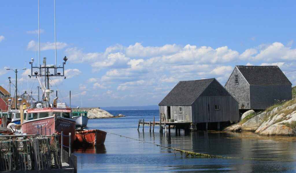 Fishing boats docked beside wooden lobster traps in a small harbor, with weathered wooden buildings on stilts over the water and a clear blue sky in the background.
