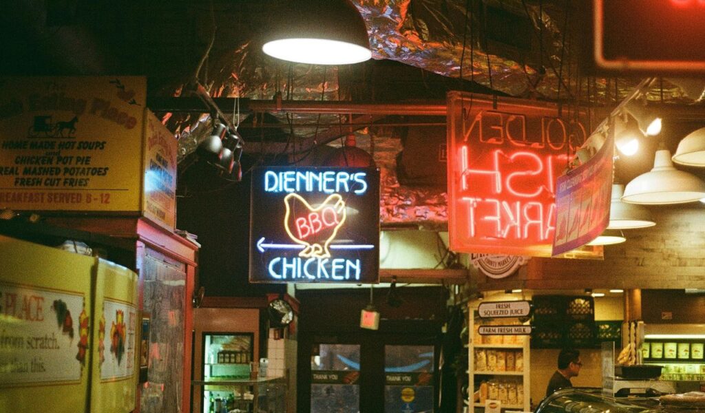 Market in Philadelphia with neon BBQ chicken and fish market signs, plus shelves of fresh juice and milk.