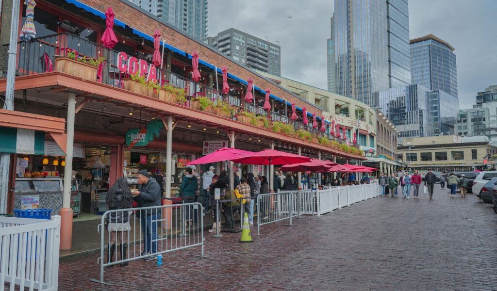 People walking and dining under red umbrellas at Pike Place Market in Seattle, with high-rise buildings in the background.