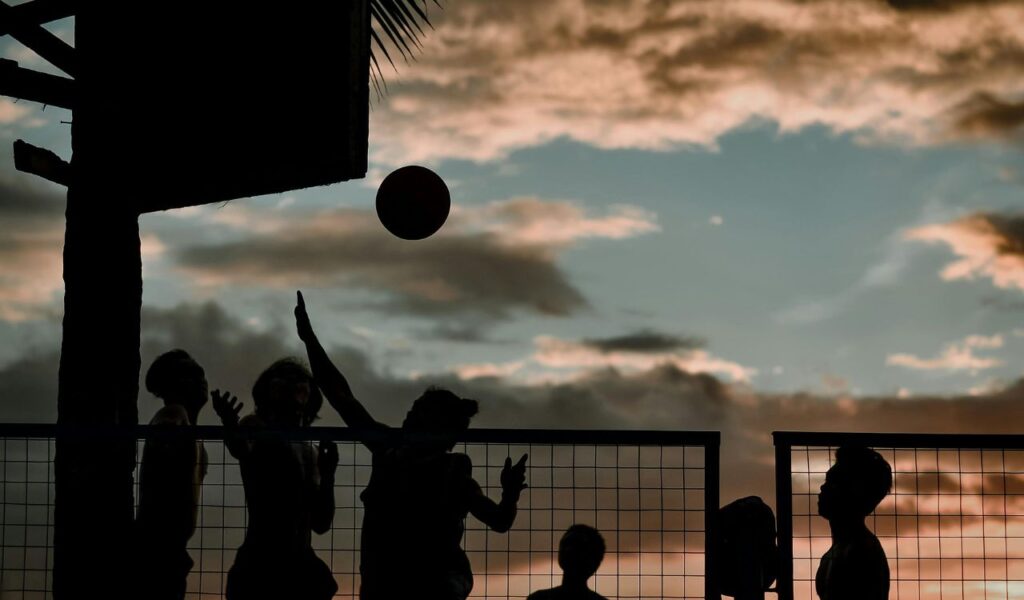 Silhouettes of people playing basketball at sunset, with the ball in midair against a cloudy sky.