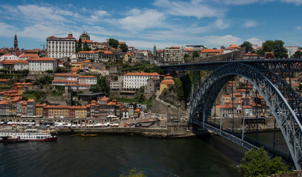 Colorful hillside buildings and red rooftops line the Douro River in Porto, Portugal, with the Dom Luís I Bridge arching overhead.