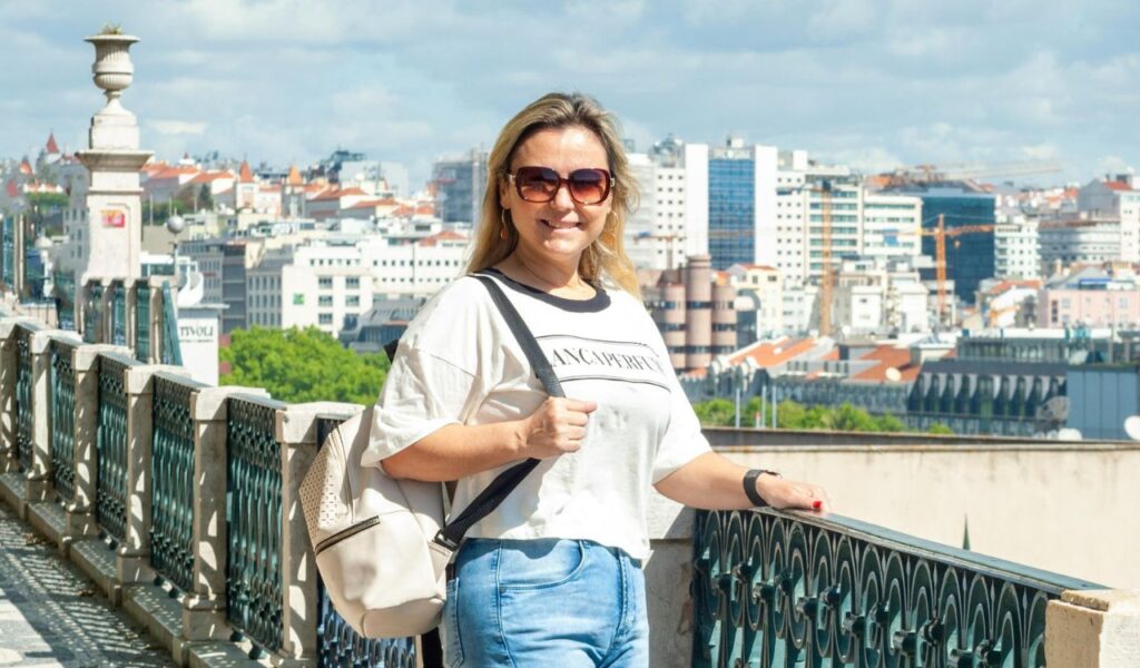 Woman in sunglasses and casual outfit stands by a railing with a sunny cityscape in the background.
