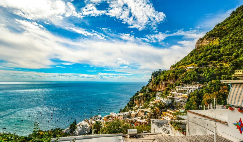 Colorful cliffside village of Positano, Italy, overlooking the sparkling blue waters of the Amalfi Coast under a bright, partly cloudy sky.
