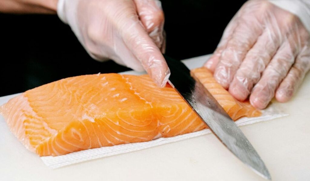 A chef wearing gloves slices raw salmon on a cutting board, demonstrating safe food handling to prevent cross-contamination.