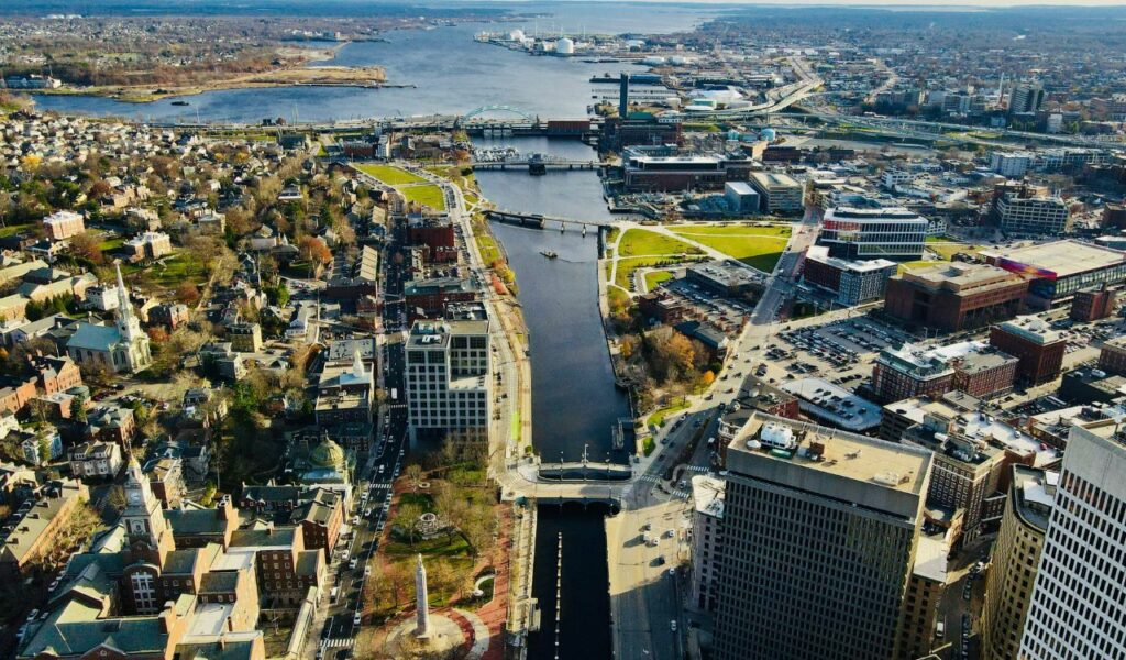 Aerial view of downtown Providence, Rhode Island, showing the Woonasquatucket River winding through the city, surrounded by historic buildings, modern high-rises, bridges, and green spaces, with Narragansett Bay visible in the distance.