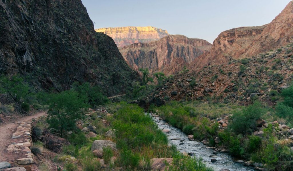 A narrow stream flows between rocky canyon walls in Pueblo, Colorado, with green vegetation lining the banks and rugged mountains in the background under a clear sky.