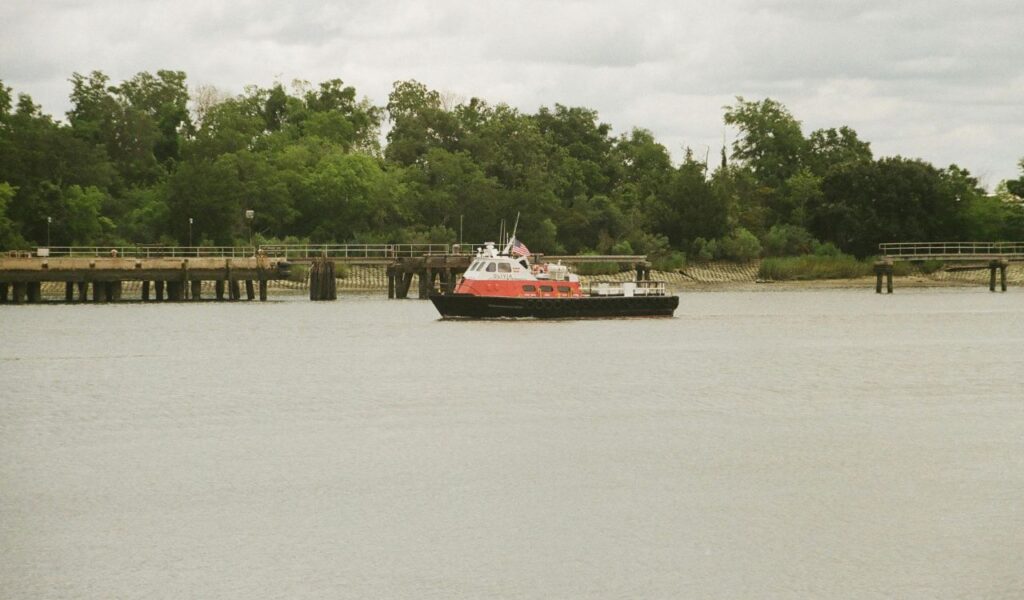 A small red and white boat with an American flag moves along the calm Savannah River, with green trees and wooden piers in the background under a cloudy sky.