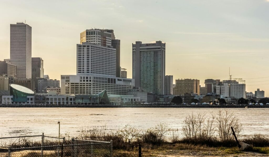  View of the New Orleans skyline from across the Mississippi River at sunset, with modern high-rise buildings, waterfront structures, and a foreground of a fence and sparse vegetation along the riverbank.