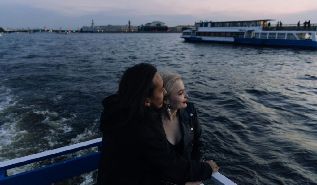 A couple embraces on the deck of a boat at sunset, with city lights and another vessel visible in the background over the rippling water.