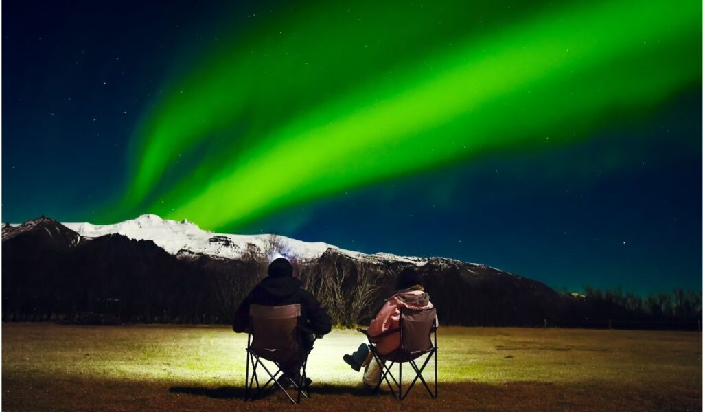 Romantic Couple in black and bright outfit sit in foldable camping chairs watch northern lights show in official campsite. Woman and man at winter night landscape in Iceland
