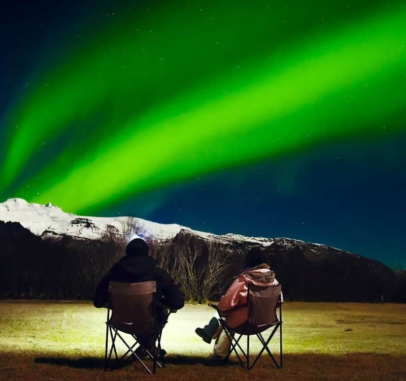 Romantic Couple in black and bright outfit sit in foldable camping chairs watch northern lights show in official campsite. Woman and man at winter night landscape in Iceland