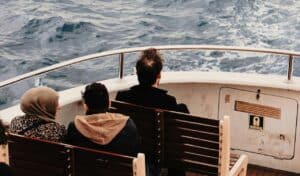 Three passengers sit on wooden benches at the back of a boat, facing the open sea with waves in the background.