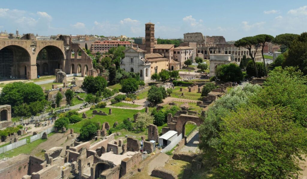 Ancient ruins of the Roman Forum in Rome with the Colosseum in the background, surrounded by historic buildings, trees, and clear blue skies.