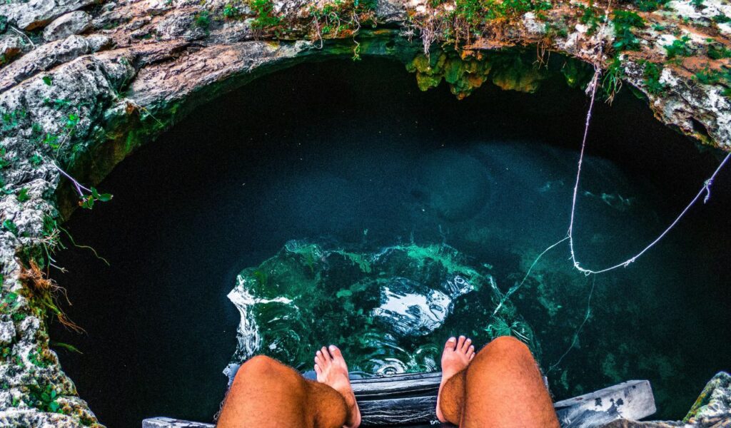 A person sitting at the edge of the Sac Actun cenote in Mexico, with feet dangling above clear blue-green water surrounded by rocky walls.