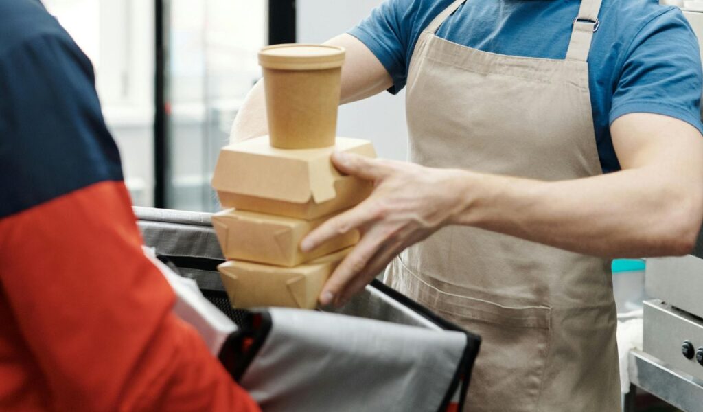 A worker in an apron places sealed food containers and a cup into an insulated delivery bag, ensuring proper storage and safe transport.