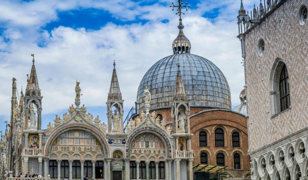 Saint Mark's Basilica in Venice featuring ornate Gothic arches, marble details, and a large domed roof under a partly cloudy sky.