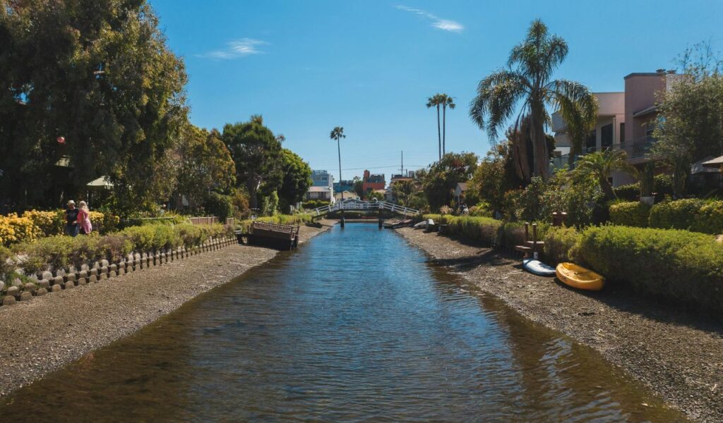 A calm waterway lined with lush greenery, palm trees, and colorful houses, with a small footbridge in the distance under a clear blue sky.

