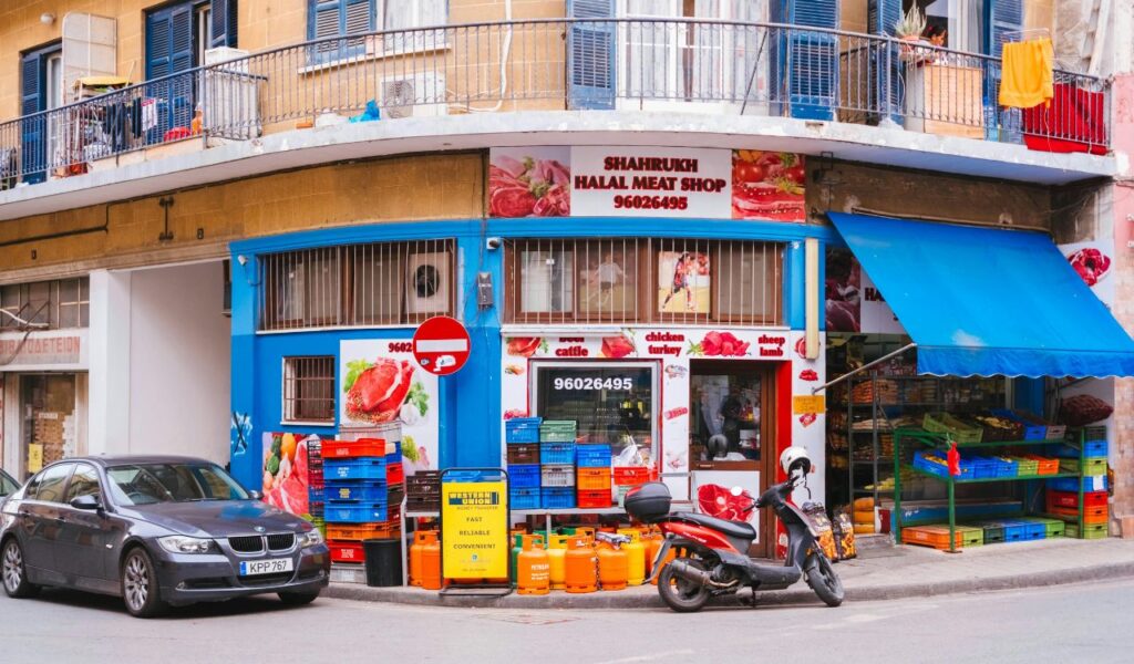 A street view of Shahrukh Halal Meat Shop with a bright blue exterior, colorful crates, propane tanks, and a parked scooter in front.