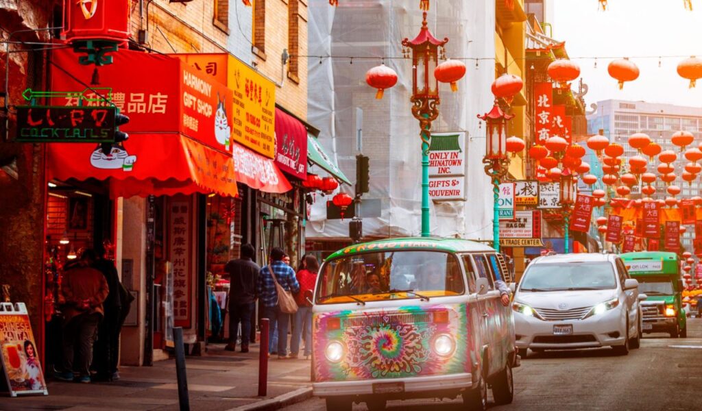 A colorful street scene in San Francisco’s Chinatown, featuring red lanterns strung across the road, vibrant shop signs, and a tie-dye painted Volkswagen van driving past pedestrians and cars.