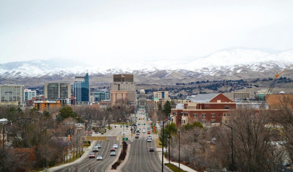 A view of downtown Boise, Idaho, with a broad avenue leading toward the city center, flanked by modern and historic buildings. Snow-covered mountains form a dramatic backdrop behind the skyline.