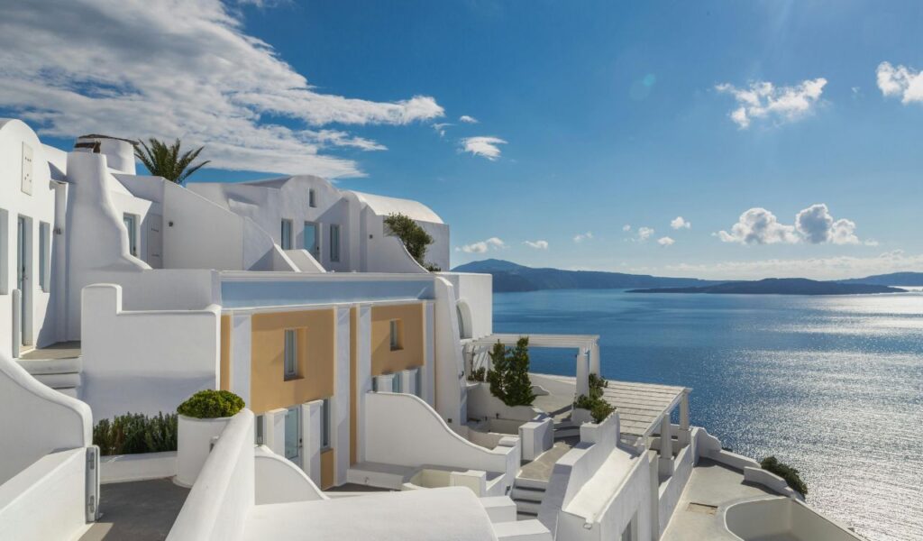 Whitewashed buildings overlooking the deep blue Aegean Sea in Santorini, Greece, under a bright sky with scattered clouds.