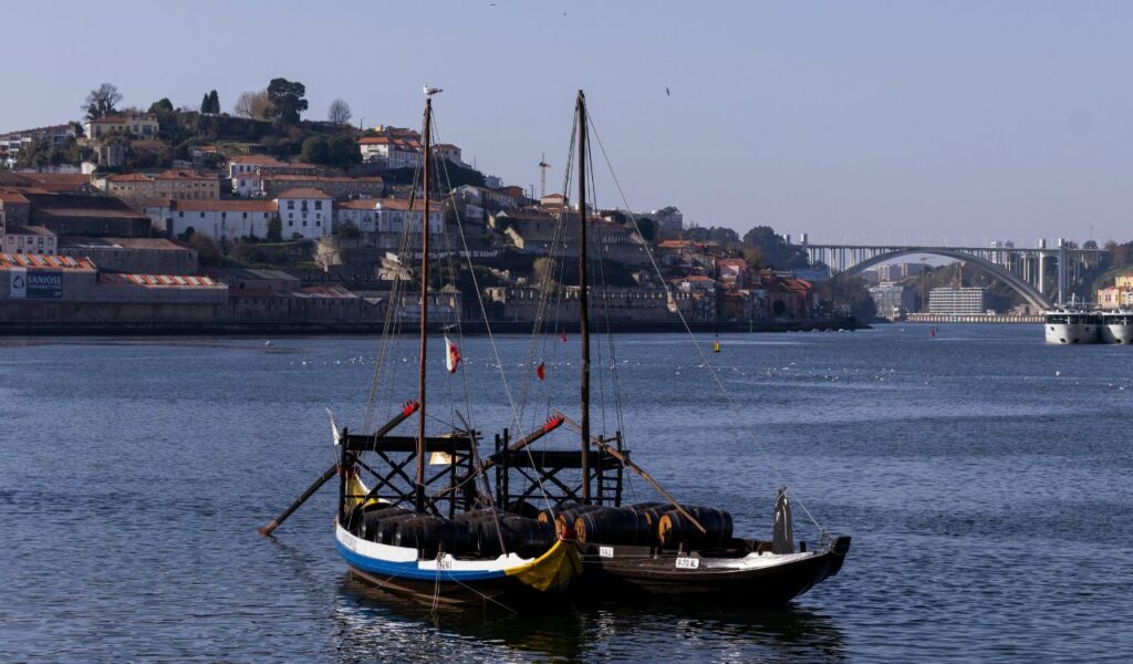 Wooden boats with wine barrels on the Douro River, Vila Nova de Gaia and Arrábida Bridge in the background.