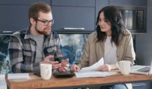 Man and woman sitting at a table discussing documents, with coffee mugs, a notebook, and a laptop in front of them.