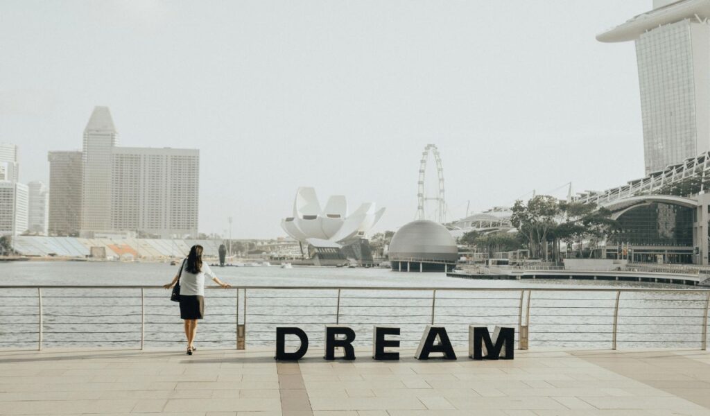 Woman stands by the waterfront in Singapore with "DREAM" letters in the foreground and the city skyline behind.