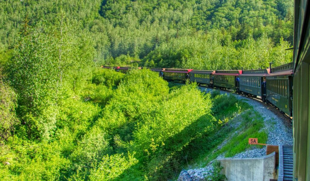 A historic White Pass and Yukon Route Railway train winds through lush green forested mountains near Skagway, Alaska, under bright summer sunlight.
