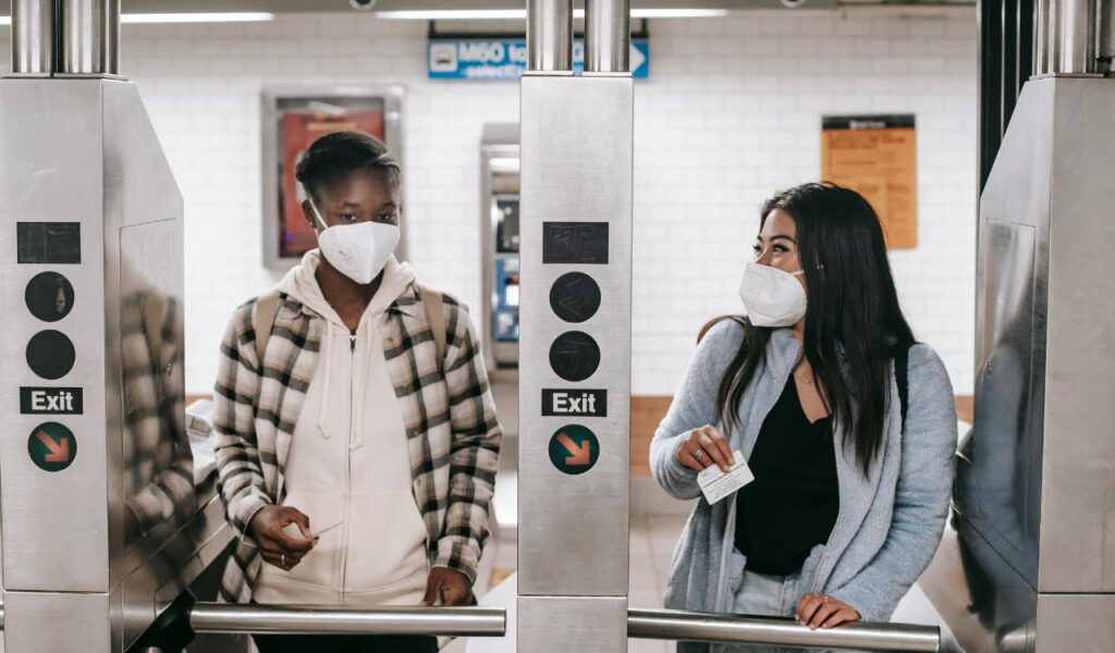 Two women wearing face masks pass through subway turnstiles, one holding a transit card, in an underground station.