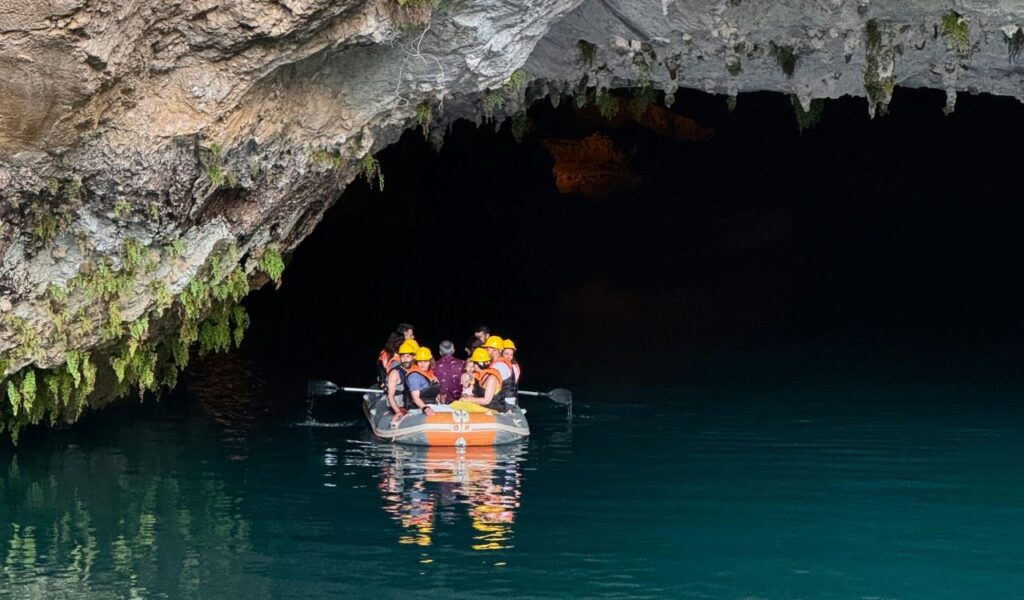 Group of people wearing helmets and life vests on a raft exploring Slovenia’s Karst caves with dark waters and rocky cave walls above
