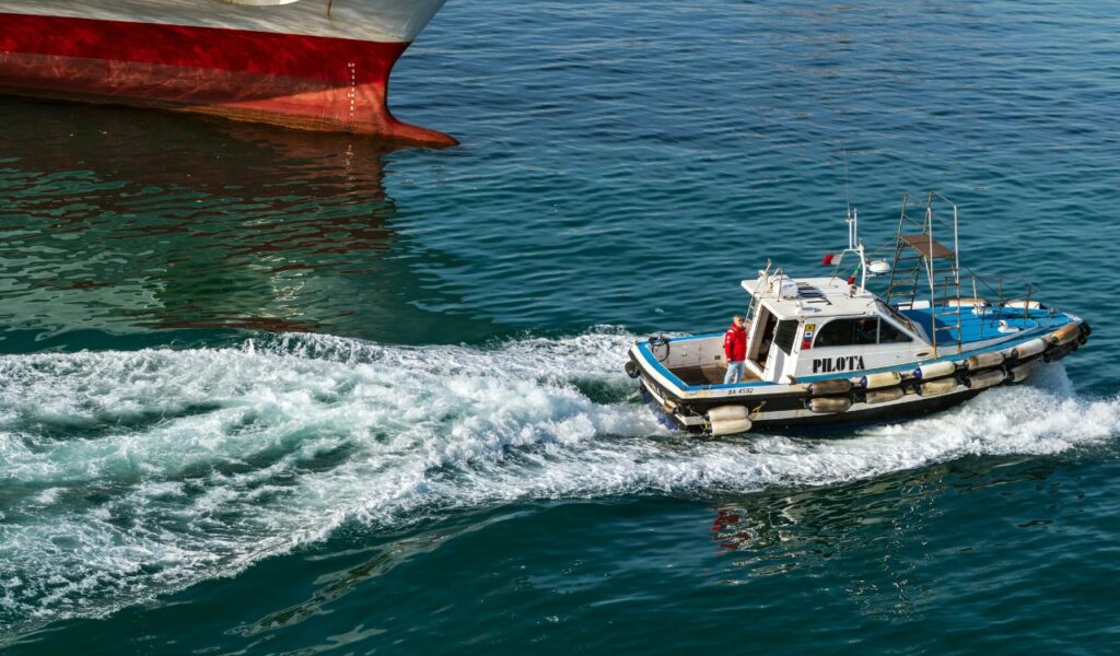 A small pilot boat labeled 'PILOTA' creating a wake as it navigates through the harbor beside a large ship's hull, with a crew member in a red jacket standing on deck