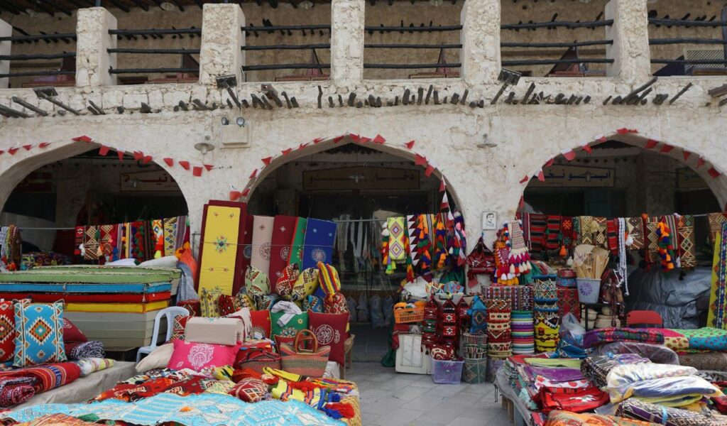  Souq Waqif market in Doha displaying colorful textiles, cushions, and traditional crafts under stone archways.