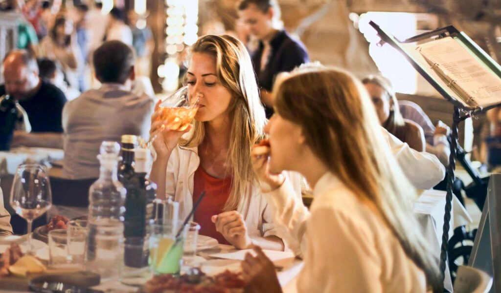 Two women enjoying food and drinks at a lively restaurant, seated at a table with glasses, plates, and condiments in a warmly lit dining atmosphere