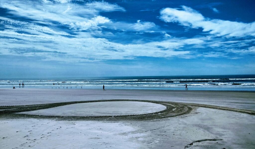 Wide sandy beach in St. Augustine, Florida, with ocean waves, a few people walking along the shore, and a circular tire track in the foreground under a partly cloudy blue sky.