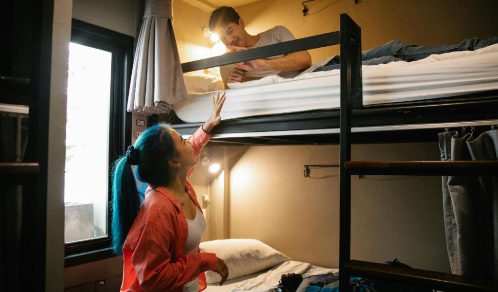 Woman in a coral shirt talking to a man lying on the top bunk bed in a cozy hostel room, with backpacks and travel gear nearby.